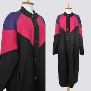 Può includere: Un cappotto lungo caratterizzato da un design a blocchi di colore nero, rosa acceso e viola. Il cappotto presenta un colletto alto, una chiusura frontale con bottoni neri e due tasche laterali. Il capo è esposto su un manichino.
