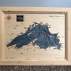 May include: A framed wooden bathymetric map of Lake Superior featuring layered blue cutouts representing water depth. The light wood frame surrounds a map with printed text, a compass rose, and coordinates reading Latitude 47.723067 and Longitude -88.940720.
