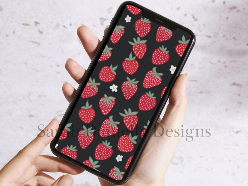 May include: A black phone case with a pattern of red strawberries and white flowers. The phone case is being held in a person's hand.
