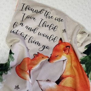 May include: A beige baby blanket featuring a watercolour illustration of two foxes. The blanket has the text "I found the one I love. I held on and would not let him go." and "Song of Solomon 3:4" printed on it.