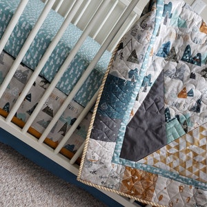 May include: A baby crib with a blue and white patterned crib sheet and a multi-colored quilt with a mountain and arrow design. The quilt has a yellow, blue, and gray color scheme. The text "Adventure Awaits" is visible on the quilt.