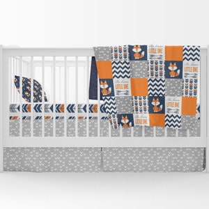May include: A white cot with a grey and orange fox themed bedding set. The bedding includes a quilt, fitted sheet, and a cot skirt. The quilt has a patchwork design with orange, grey, and navy blue squares. The fitted sheet has a pattern of orange and navy blue arrows. The cot skirt has a grey and white pattern of small hearts. The quilt features the text "Be Brave Little One" repeated throughout.