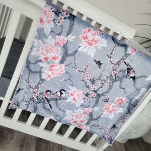 May include: A gray and white baby blanket with a floral pattern featuring pink flowers, branches, and birds. The blanket has a soft, plush texture.