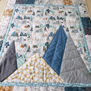 May include: A white and grey baby quilt with a mountain theme. The quilt features three large mountains made of different fabrics, including blue, grey, and yellow. The mountains are surrounded by smaller mountains and the text "Adventure Awaits".