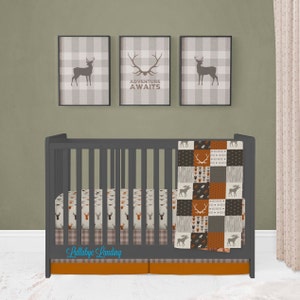 Puede incluir: Una cuna gris con una colcha de retazos marrón y beige y una sábana bajera con estampado de ciervo. La cuna tiene una falda a cuadros gris y blanca y un volante naranja. Tres marcos con siluetas de ciervos y el texto "La aventura te espera" están en la pared detrás de la cuna.