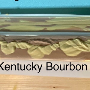May include: Two bars of handmade soap in clear plastic molds. The soap is a swirl of brown and yellow. The bottom bar has the text "Kentucky Bourbon" printed on the mold.