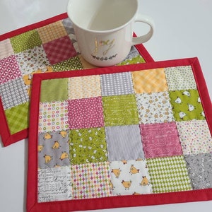 May include: Two patchwork placemats with a red border, featuring squares of various patterned fabrics in green, pink, yellow, and gray. A white mug with gold lettering sits on one. The fabrics include floral, chicken, and text patterns.