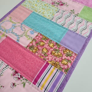 May include: A patchwork table runner with a purple border. The runner is made of various pink, green, blue, and yellow fabrics with floral and geometric patterns.