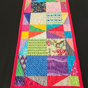 May include: A colorful patchwork table runner with a variety of geometric and floral patterns. The runner is trimmed with a bright pink border.