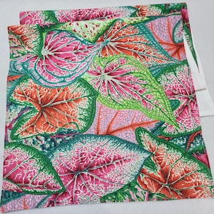May include: A folded fabric featuring a vibrant botanical pattern. The design showcases large, detailed leaves in shades of pink, green, and orange, with intricate veining. The fabric has a white edge, suggesting it may be a tablecloth or other textile.