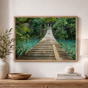 May include: A rectangular wall art print featuring a wooden suspension bridge stretching across a vibrant turquoise river, surrounded by dense green forest foliage. The print is displayed in a light-toned wooden frame on a neutral-colored wall.