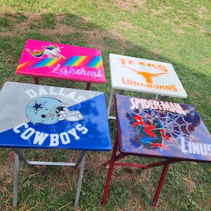 May include: Four folding tables with painted designs. One table features a unicorn and the name "Lakshmi", another displays the Dallas Cowboys logo, a third shows the Texas Longhorns logo, and the last has a Spider-Man graphic and the name "Linus".