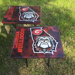 以下が含まれることがあります： 赤と黒の大理石模様の小さな正方形のテーブルが2つ。各テーブルには、ジョージアブルドッグスのロゴとオレンジ色の「GEORGIA BULLDOGS」の文字が描かれています。テーブルは黒い金属製の脚で、草の生えた表面に置かれています。
