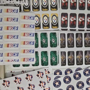May include: A collection of stickers featuring various sports teams and racing logos. Includes NASCAR, Pittsburgh Steelers, Kansas City Chiefs, Dallas Stars, New Orleans Saints, and Houston Astros designs. The stickers are in various shapes and sizes.