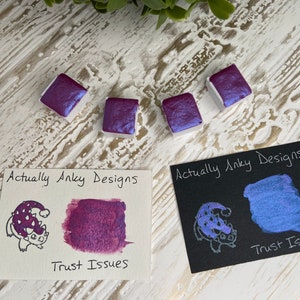 May include: Four square watercolor paint pans in shades of purple and blue with a pearlescent sheen. Two cards display swatches of the paint, one on white and one on black, with the text "Actually Anky Designs" and "Trust Issues".