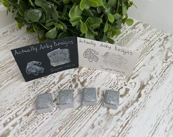May include: Four silver glitter watercolor paint pans with the text "Actually Anky Designs" and "Supernova" on two black and white cards.