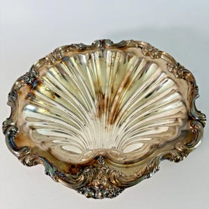 May include: A silver-plated serving dish shaped like a large scallop shell. The bowl features deep, radiating fluted grooves and a wide, ornate border with embossed scrollwork and floral details. The metal surface shows areas of natural tarnish.