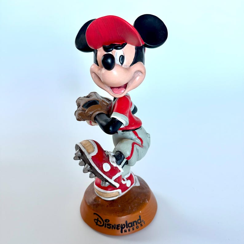 Peut inclure: Figurine de Mickey Mouse en uniforme de baseball, pr&ecirc;t &agrave; lancer la balle. Il porte une casquette, un maillot et des chaussures rouges, un pantalon gris et un gant marron. La base est marron avec le logo Disneyland Resort.