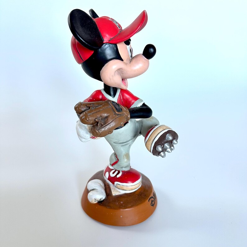Peut inclure: Figurine de Mickey Mouse en uniforme de baseball. Mickey porte une casquette, un maillot et des chaussures rouges, avec un pantalon gris et un gant marron. La figurine est sur une base marron.