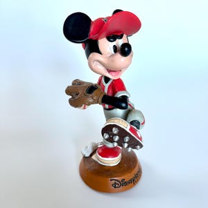 Peut inclure: Figurine de Mickey Mouse en uniforme de baseball, pr&ecirc;t &agrave; lancer. Mickey porte une casquette et un maillot rouges avec des d&eacute;tails noirs, un gant marron et des crampons rouges et blancs. La base est marron et porte le texte "Disney Parks".
