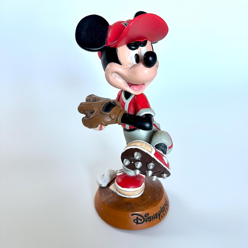Peut inclure: Figurine de Mickey Mouse en uniforme de baseball, pr&ecirc;t &agrave; lancer. Mickey porte une casquette et un maillot rouges avec des d&eacute;tails noirs, un gant marron et des crampons rouges et blancs. La base est marron et porte le texte "Disney Parks".