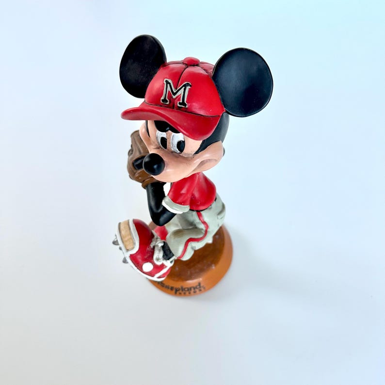 Peut inclure: Figurine de Mickey Mouse en uniforme de baseball, portant une casquette rouge avec un "M" noir. La figurine tient un gant de baseball et est pos&eacute;e comme pr&ecirc;te &agrave; lancer. La base est marron avec "Disneyland" imprim&eacute;.