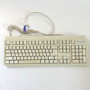 May include: A beige Packard Bell computer keyboard with a coiled cable and a purple PS/2 connector. The keyboard features a standard QWERTY layout, a numeric keypad, function keys, and a spacebar with printed text regarding the Quick Start Guide.