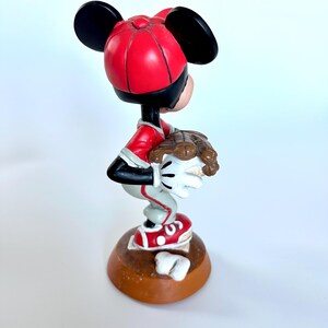Peut inclure: Figurine de Mickey Mouse en uniforme de baseball, avec une casquette et un maillot rouges, un pantalon gris et des chaussures rouges. La figurine tient un gant de baseball et se tient sur une base marron.