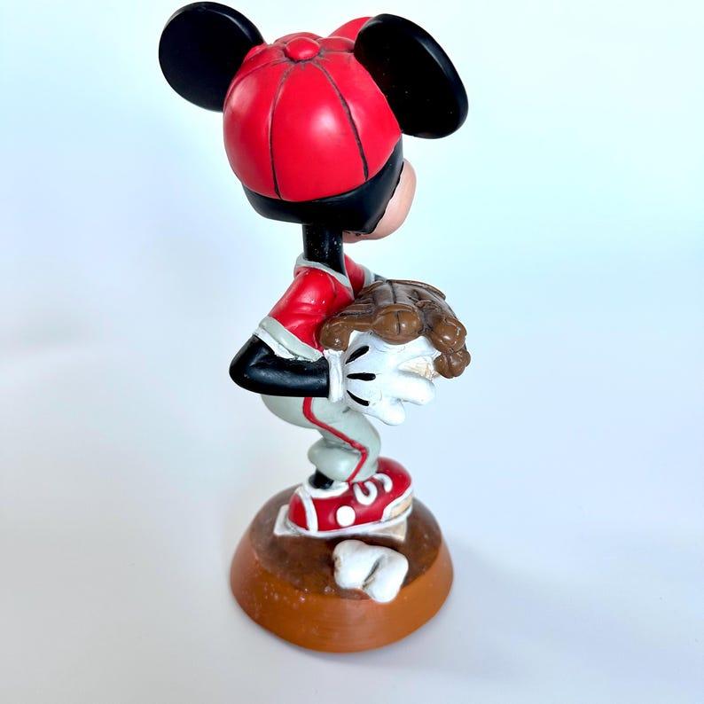 Peut inclure: Figurine de Mickey Mouse en uniforme de baseball, avec une casquette et un maillot rouges, un pantalon gris et des chaussures rouges. La figurine tient un gant de baseball et se tient sur une base marron.