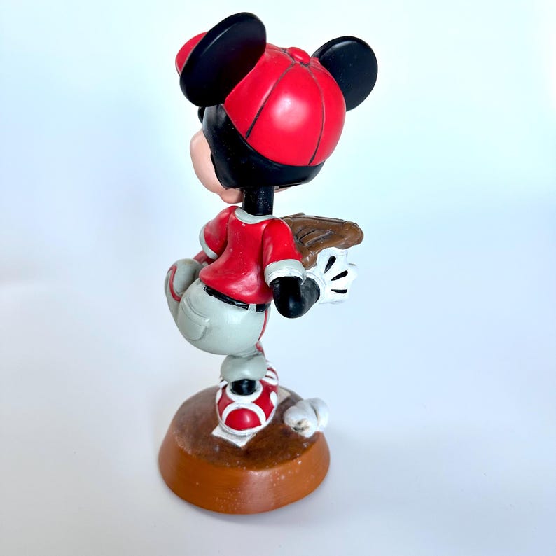 Peut inclure: Figurine d'un personnage de dessin anim&eacute; en uniforme de baseball. La figurine porte une casquette, un maillot et des chaussures rouges, avec un pantalon gris. Elle tient un gant de baseball et se tient sur une base marron.