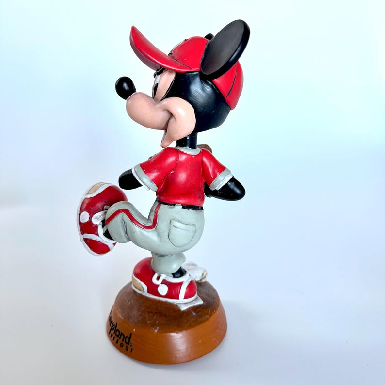 Peut inclure: Figurine de Mickey Mouse en uniforme de baseball. Mickey porte une casquette, un maillot et des chaussures rouges, avec un pantalon gris. La figurine est sur une base marron avec le texte "Cleveland Resort".
