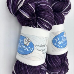 May include: Two skeins of hand-dyed yarn in shades of deep purple, lavender, and white. The yarn is labeled with the text "Ladyfernaco Creations" and "Fun, fanciful yarn, Hand dyed with Love". The yarn is tightly wound and ready for crafting.