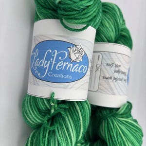 May include: Two skeins of green yarn with white accents. The yarn is labeled "Lady Fernaco Creations" on a blue and white label. The yarn is a vibrant green color, perfect for knitting or crocheting projects.
