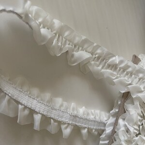 May include: White elastic trim with a ruffled edge. The trim is made of a soft, delicate fabric and has a subtle sheen.