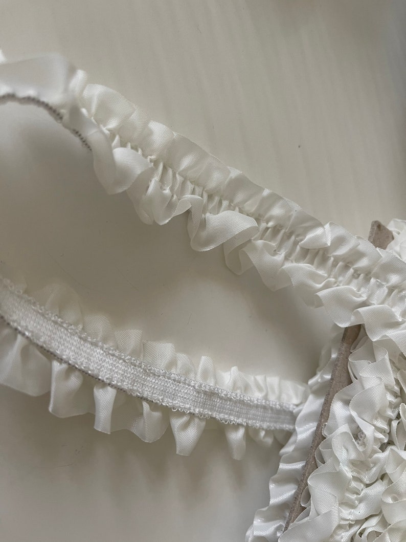 May include: White elastic trim with a ruffled edge. The trim is made of a soft, delicate fabric and has a subtle sheen.