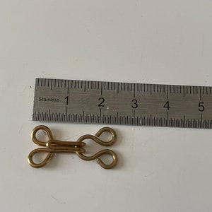 Op de afbeelding: Een gouden metalen haak- en oogsluiting met een lengte van ongeveer 2,5 cm. De sluiting is bedoeld om op stof te worden genaaid.
