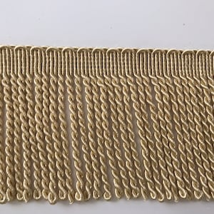Op de afbeelding: Een close-up van een beige gedraaide franje trim. De franje is gemaakt van een zacht, geweven materiaal en heeft een subtiele glans.