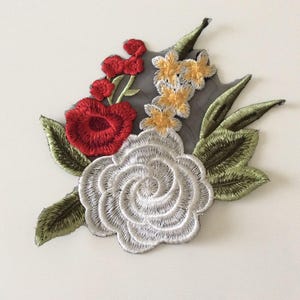 Peut inclure: Écusson floral brodé avec une grande rose blanche, une rose rouge et de petites fleurs jaunes et rouges. Des feuilles vertes entourent les fleurs, le tout sur un support gris. Ce patch décoratif est idéal pour ajouter une touche d'élégance aux vêtements ou accessoires.
