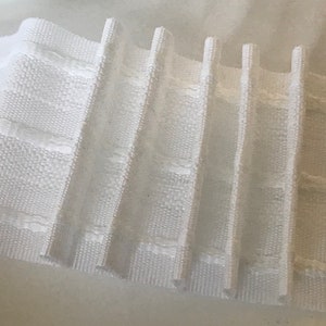 Könnte beinhalten: Weißes, gefaltetes Gardinenband mit einem Gittermuster. Das Band ist in Falten gelegt, wodurch ein dekoratives und funktionales Design für Gardinen entsteht.