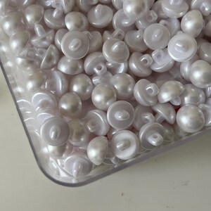 Pode incluir: Um close-up de um recipiente cheio de botões brancos perolados. Os botões têm um acabamento brilhante e estão dispostos em um padrão aleatório.