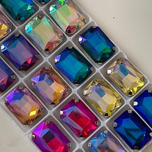 Può includere: Una varietà di strass rettangolari iridescenti in un vassoio di imballaggio in plastica. Gli strass sono disposti in file e colonne, e sono tutti di colori diversi, tra cui blu, verde, viola e rosa.