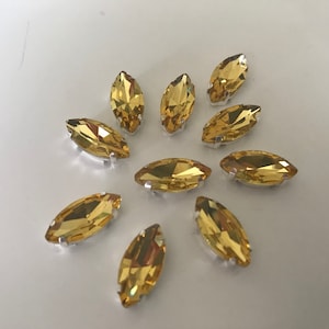Puede incluir: Diez piedras de imitación de forma marquesa de color dorado con engastes plateados. Las piedras de imitación están dispuestas en un patrón circular sobre un fondo blanco.