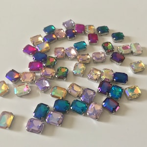May include: Assortment of iridescent rectangular rhinestones in various colors, including pink, purple, blue, green, and yellow. The rhinestones are scattered on a white surface.