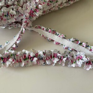 May include: White elastic trim with a pink and blue floral pattern. The trim has a ruffled edge and is perfect for adding a touch of elegance to any project.