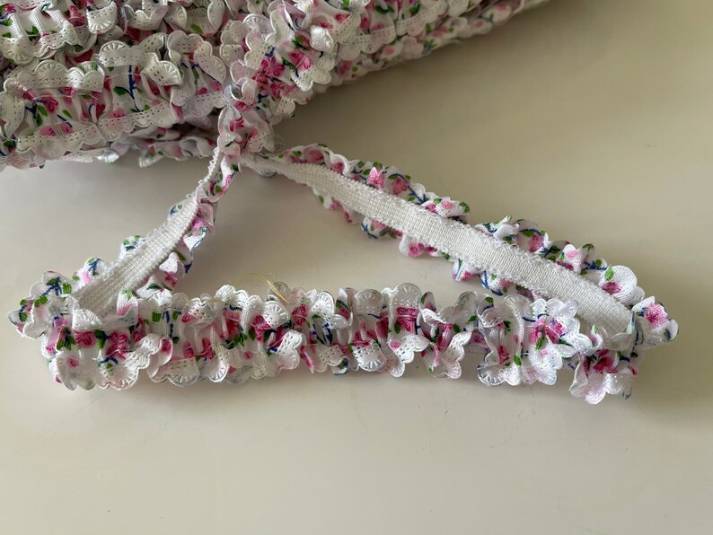 May include: White elastic trim with a pink and blue floral pattern. The trim has a ruffled edge and is perfect for adding a touch of elegance to any project.