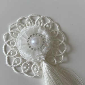 Peut inclure: Garniture en dentelle blanche avec une fleur blanche et une perle au centre. La fleur est ornée de strass autour de la perle. Un pompon blanc pend en bas.
