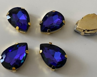 Könnte beinhalten: Vier tropfenförmige, dunkelblaue Glas-Straßsteine mit goldenen Fassungen. Die Strasssteine haben ein facettierten Design und eine Hochglanz-Oberfläche.