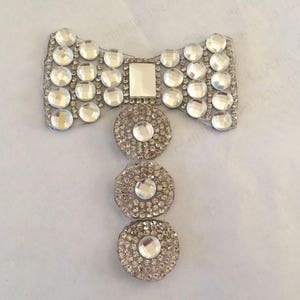 Puede incluir: Un adorno de pedrería plateado en forma de lazo con tres pedrerías redondas unidas a la parte inferior.