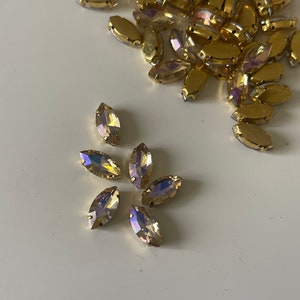 Peut inclure: Un groupe de strass de forme marquise ton or avec des facettes irisées. Les strass sont éparpillés sur une surface blanche.