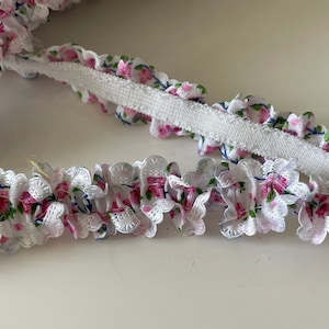 May include: White elastic trim with a pink and blue floral print. The trim has a ruffled edge and is perfect for adding a touch of elegance to any project.